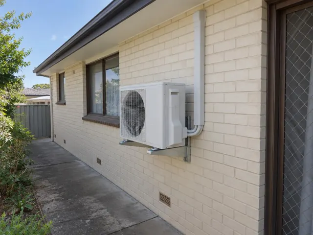 Outdoor air conditioner condenser on a brick house wall beside windows and a driveway in Gilles Plains