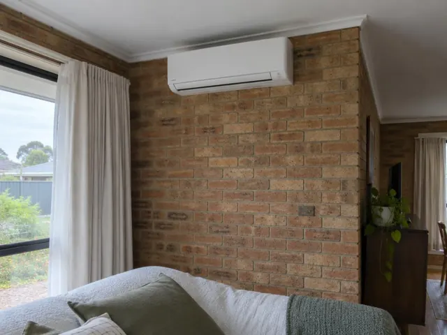 Wall-mounted split system above a brick feature wall in a living room with curtains and a window in Ingle Farm